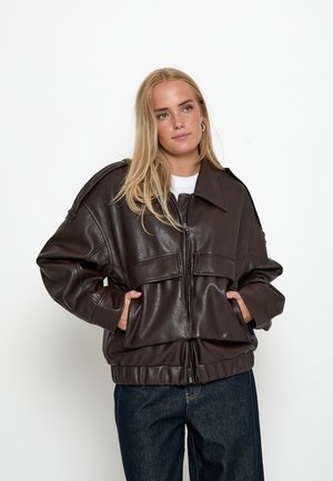 Junge Frau mit blondem Haar, die eine dunkelbraune übergroße Lederjacke und dunkelblaue Jeans trägt, Hände in den Jackentaschen, neutraler Hintergrund.