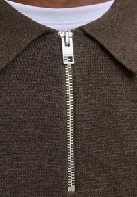 Detailní pohled na hnědý pletený svetr se stříbrným zipem, který je částečně rozepnutý přes bílý tričko s kulatým výstřihem.