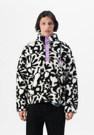 Mustavalkoinen kukkakuvioinen fleece-takki, jossa on korkea kaulus, violetti napitus ja vetoketjullinen tasku. Teksturoitu kangas rentoa istuvuutta varten.