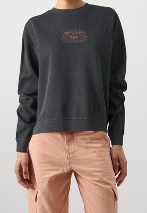 Persona che indossa una felpa nera a maniche lunghe con una piccola grafica e pantaloni cargo rosa chiaro, in piedi su uno sfondo neutro.