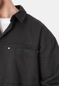 Chemise en denim noir avec col boutonné, poche poitrine avec logo de la marque en relief et détails de couture visibles.