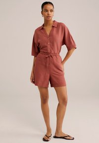 Rödbrun linne kort jumpsuit med krage, knäppning fram, lösa ärmar, dragsko i midjan och fickor. Bärs med svarta flip-flops.