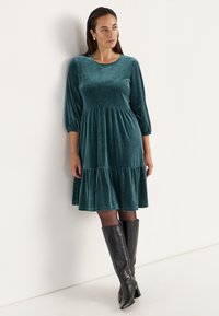 Turquoise fluwelen jurk met een ronde halslijn, driekwartmouwen en een uitlopende tiered rok. Gepaard met zwarte laarzen tot de knie.