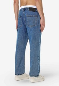 Siniset rentot istuvaiset farkut, joissa on kaksi takataskua ja musta nahkainen merkkilaatta vyötäröllä. Valkoiset tennarit näkyvissä.