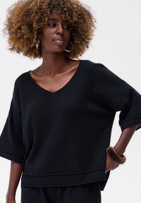 Vrouw met krullend haar draagt een zwarte V-hals textuurtop en bijpassende broek, accessorized met oorringen en een bontarmband om haar pols.