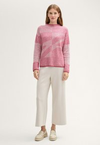Strickpullover in Rosatönen mit geometrischen Mustern, gerippter Textur und hohem Kragen, kombiniert mit cremefarbenen weit geschnittenen Hosen und beigen Plateauschuhen.