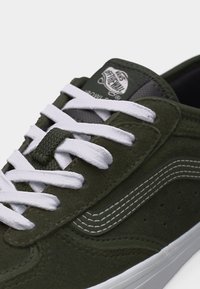 Temno zelena semiš tekaška copata Vans z belimi vezalkami, belim bočnim trakom in logotipom Vans "Off The Wall" na jeziku.