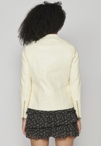 Veste en cuir couleur crème avec des accents zippés, design ajusté et surface texturée. Présente des épaules structurées et des coutures verticales.