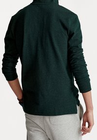 Dunkelgrüner Pullover, strukturierter Stoff, lange Ärmel, hoher Kragen, lockere Passform, getragen mit grauen Jogginghosen, sichtbare Manschettendetails.