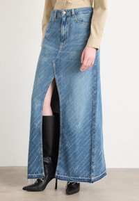 Denim maxikjol med framklippning som visar svarta högklackade stövlar, matchad med en beige långärmad topp, står på en enkel golv.