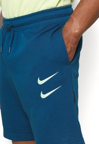 Teal Nike shorts featuring a drawstring waistband, side pockets, and two contrasting swoosh logos in light green. Soft cotton blend material.