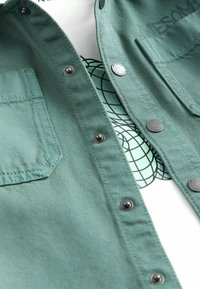 Chemise en denim verte avec deux poches poitrine et boutons-pression argentés, superposée à un t-shirt blanc à motif géométrique.
