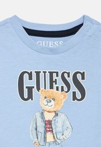 Ανοιχτό μπλε βαμβακερό t-shirt με γραφικό ενός αρκούδου σε τζιν μπουφάν, τυπωμένο το κείμενο "GUESS" σε μαύρο, με απαλή υφή.
