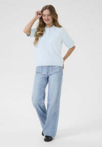 Himmelblaues Polo mit kurzen Ärmeln und strukturiertem Muster, kombiniert mit weiten, hellblauen Jeans. Schwarze Schuhe runden das Outfit ab.