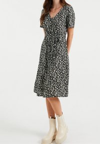 Czarna sukienka w kwiaty z krótkimi rękawami, dekoltem w serek, wcięciem w talii oraz spódnicą sięgającą kolan; zestawiona z beżowymi, grubymi botkami za kostkę.