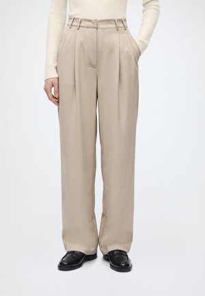 Modèle portant un pantalon beige ample à pinces avec des passants de ceinture, un pull côtelé couleur crème et des mocassins noirs, sur un fond clair uni.