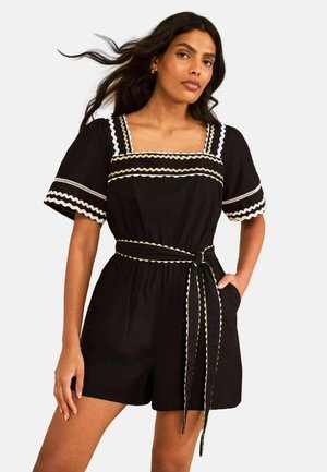 Mujer que lleva un mono corto negro con ribete beige de zigzag, mangas cortas abullonadas, escote cuadrado y cinturón atado a la cintura.