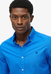 Blauw overhemd met knoopsluiting en een kraag, voorzien van een klein geborduurd logo op de borst. Glad katoen en gestructureerd ontwerp.