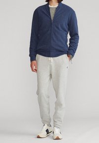 Giacca a zip blu navy con colletto alto e tasche laterali, abbinata a pantaloni della tuta grigi chiari. Sneakers bianche con accenti blu navy completano l'outfit.