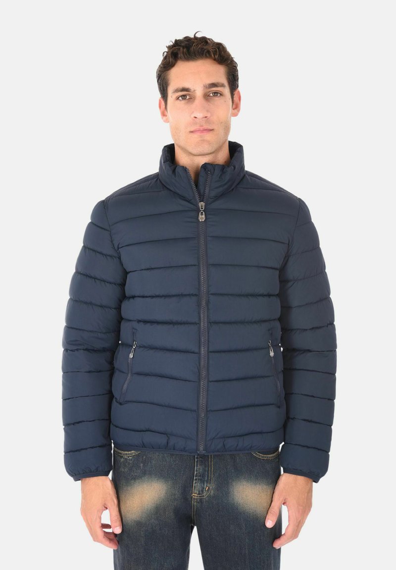 Giacca a vento blu scuro con colletto alto, design trapuntato orizzontale, zip frontale e tasche laterali. Indossata sopra jeans azzurri.