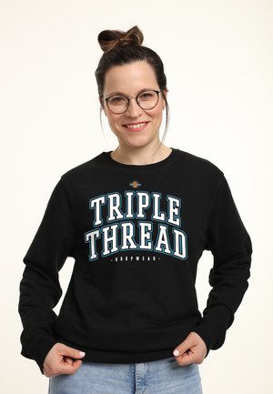 Lächelnde Frau mit Brille und Dutt, trägt ein schwarzes Sweatshirt mit der Aufschrift "TRIPLE THREAD HOOPWEAR" und blaue Jeans.