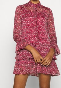 Floral-printed dress in sheer fabric, featuring a high neck, long sleeves, tiered ruffles, and shades of red, pink, and cream.