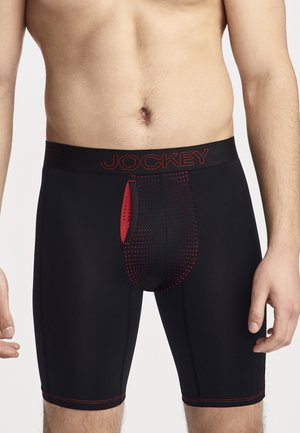 Männlicher Torso trägt schwarze und rote Jockey Boxershorts mit einem gemusterten Frontpanel und Logo-Bund, steht vor weißem Hintergrund.