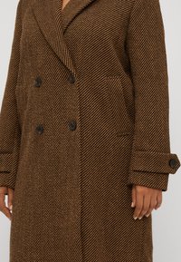 Cappotto doppiopetto in motivo herringbone marrone, con bottoni neri, ampi risvolti e tasche laterali. Tessuto testurizzato con una forma strutturata.