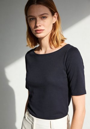 Young woman with light brown hair wearing a black short-sleeve shirt and white pants, standing against a plain light background.