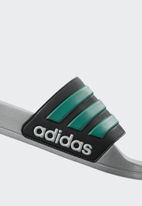 Šedý adidas šľapák s čiernym pásikom, ktorý má tri tyrkysové pruhy a biely adidas logo na boku.