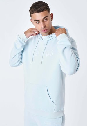 Joven ajustándose la capucha de una sudadera azul clara con texto bordado en blanco en el pecho y pantalones a juego.