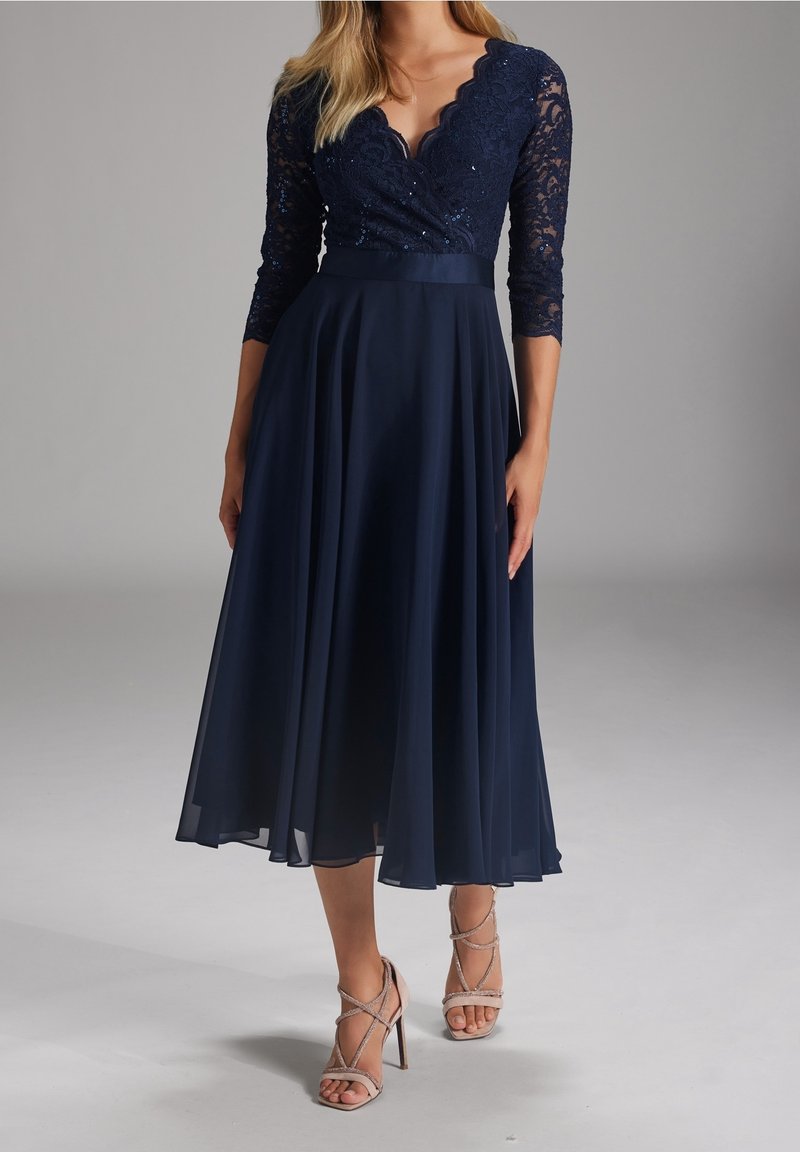 Robe bleu marine avec corsage en dentelle, manches trois-quarts et jupe fluide. Présente un décolleté en biais et des sequins subtils. Assortie à des talons à brides.