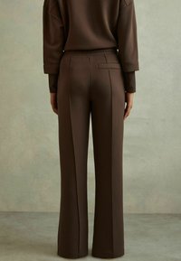 Bruine, rekbare, hoog-getailleerde broek met een rechte pijpdesign, voorzien van een enkele achterzak en naaddetails. Zachte textuur.
