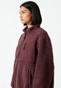 Giacca in pile bordeaux con collo alto, tasca frontale con cerniera e dettagli in tessuto liscio a contrasto. Aspetto strutturato e accogliente.