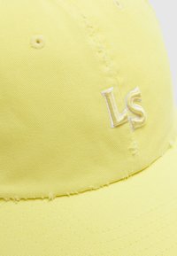 Κοντινό πλάνο ενός κίτρινου υφασμάτινου καπέλου μπέιζμπολ με ραμμένες οπές αερισμού και κεντημένα αρχικά "LS".