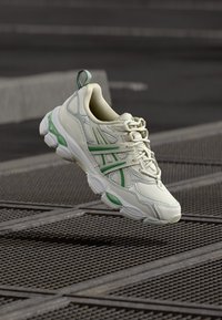 Světlá béžová sportovní obuv se zelenými detaily, texturovanou kůží a síťovinovými materiály. Má zaoblenou špičku a výrazné zavazování na tkaničky.