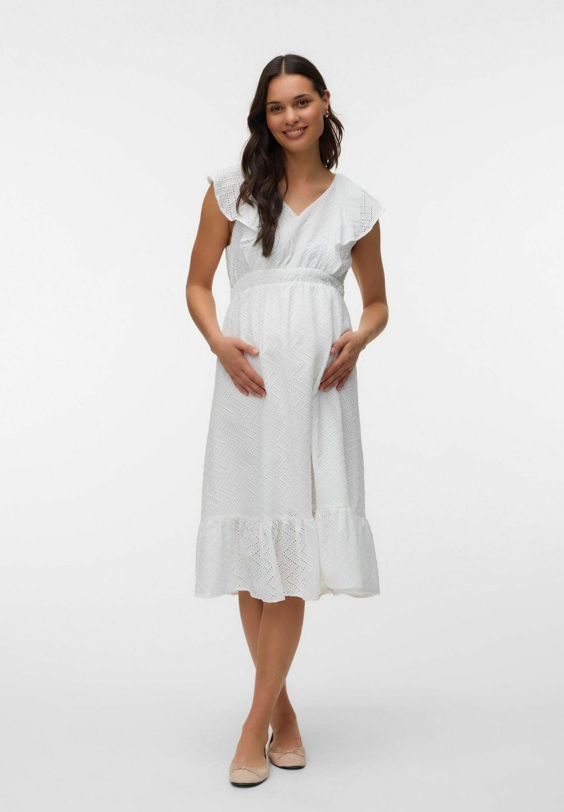 Femme enceinte en robe blanche ajourée avec manches volantées, debout pieds nus sur un fond blanc uni, souriante, les mains sur le ventre.