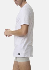 White cotton t-shirt with short sleeves, featuring a black Adidas logo on the lower left side. Model wearing gray shorts underneath.