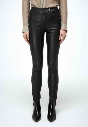 Zwart leren pantalon in slim-fit, gedragen met zwarte enkellaarsjes met puntige neus en een beige overhemd met knopen, zichtbaar bij de taille en polsen.