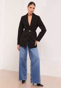Kvinde poserende i sort bælteblazer, vidde blå jeans, sorte spidse støvler og guld hoop øreringe mod en ensfarvet lys baggrund.