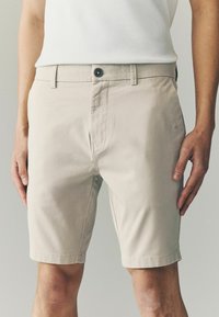 Beige shorts lavet af bomuld, med knaplukning, bæltestropper og en skræddersyet pasform, samt opkrøllede kanter og en glat tekstur.