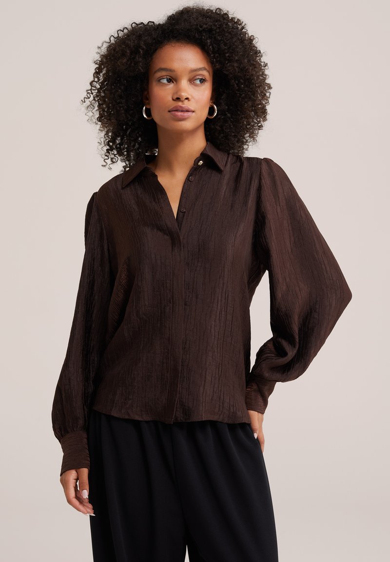 Femme aux cheveux bouclés portant un chemisier ample marron foncé texturé avec des manches longues bouffantes et un pantalon noir, regardant sur le côté.