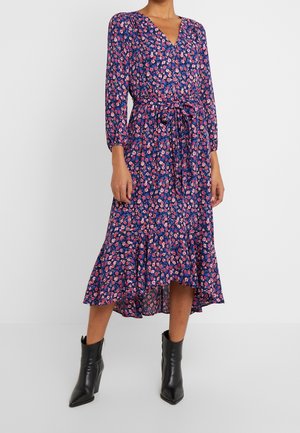 Kobieta ubrana w granatową sukienkę midi z różowym kwiatowym wzorem, z długimi rękawami, wiązaną w talii, z falbaną na dole oraz czarne botki za kostkę.