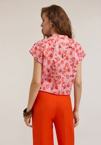 Kurzärmlige Bluse aus pinkem Stoff mit floralem Muster, kombiniert mit hochgeschnittenen orangen Hosen, die eine lockere Passform und glatte Textur zeigen.