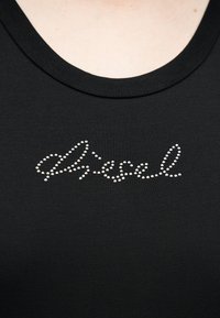 Μαύρο μπλουζάκι με στρογγυλή λαιμόκοψη και λογοτύπο με καλλιγραφικά γράμματα, σχηματισμένο από μικρές λευκές χάντρες στο κέντρο του στήθους.