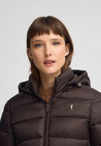 Chaqueta de plumas marrón con cierre frontal, capucha elástica y paneles cosidos. Presenta un sutil detalle de logo en el lado izquierdo del pecho.