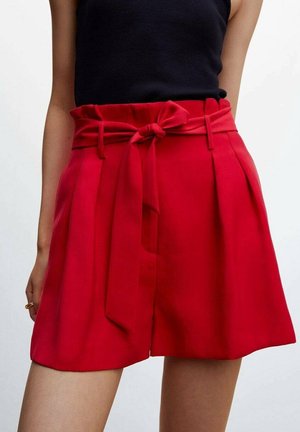 Mujer con pantalones cortos de tiro alto de color rojo brillante con cinturón anudado y pliegues, combinados con una blusa sin mangas negra, de pie contra un fondo blanco.