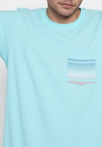 Camiseta de manga corta azul claro con un diseño de rayas texturizadas, que presenta un bolsillo frontal con rayas horizontales azules y rosas.