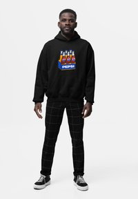 Uomo che indossa una felpa nera con cappuccio decorata con una grafica colorata di una confezione di bottiglie di Pepsi, pantaloni neri a scacchi e sneakers nere, in piedi su uno sfondo bianco.