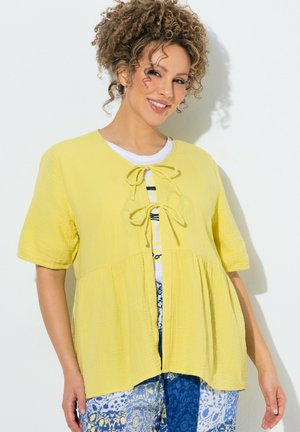 Vrouw met krullend haar die glimlacht, een gele blouse met korte mouwen die aan de voorkant is geknoopt, over een wit topje en een blauw patroon rok draagt.